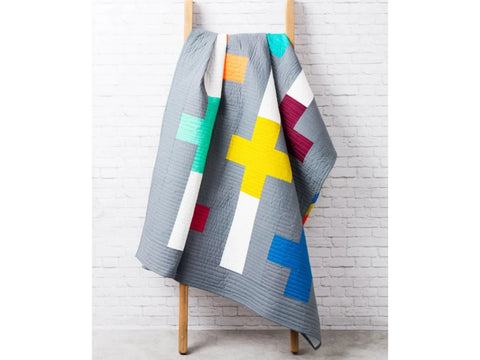 A gray quilt with colorful plus signs hangs on a wooden ladder against a white brick wall.