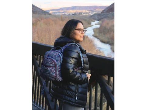 A woman wearing a black puffer jacket and glasses, with a patterned backpack, looks out over a river and valley.
