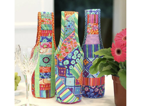 Three colorful, patchwork fabric wine totes stand on a table with a wine glass and flowers.