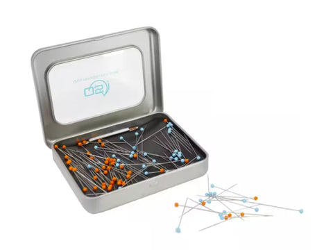 An open magnetic pin tin filled with orange and blue glass head pins, with some scattered outside.