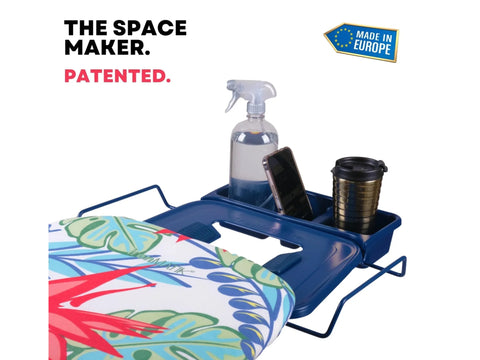 Ironing board with a tropical blue cover and a blue accessory tray holding a spray bottle, phone, and cup.
