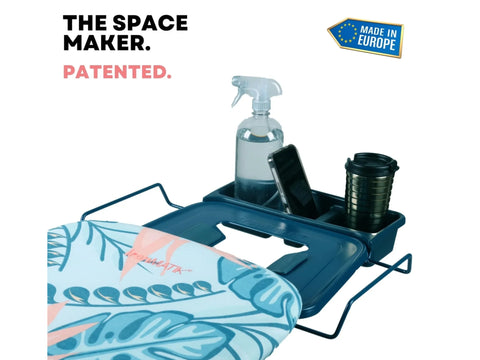 An ironing board with a tropical teal cover and a blue accessory tray holding a spray bottle, phone, and cup.