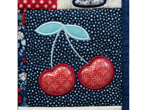 A quilted fabric block features two red cherries with light blue leaves on a navy blue star-patterned background.