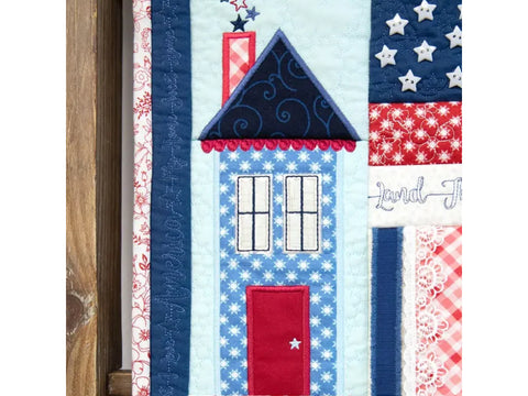 A patriotic quilted wall hanging features a house, stars, and stripes in red, white, and blue.
