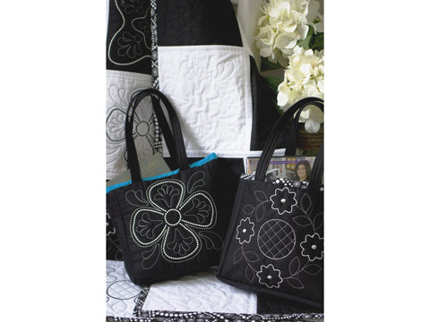 Two black tote bags with white floral embroidery sit on a black and white quilted surface.