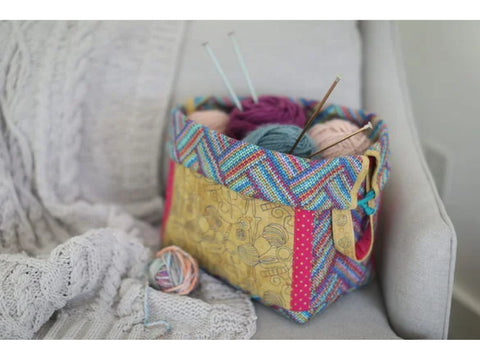 A colorful fabric knitting basket filled with yarn and needles sits on a gray blanket.