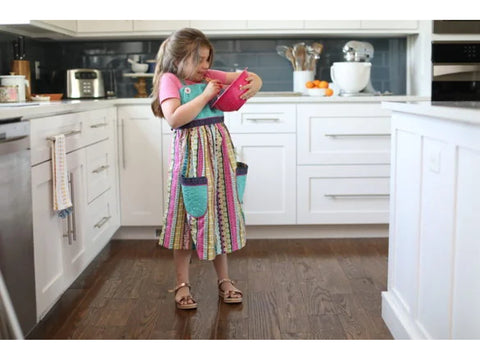 A young girl in a colorful apron tastes from a pink bowl in a modern kitchen.