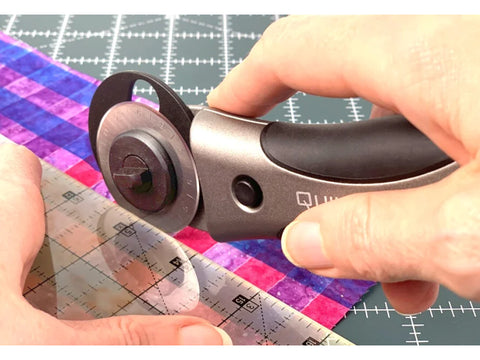 A hand holds a Quilters Select 28mm Deluxe Rotary Cutter, cutting plaid fabric on a mat.