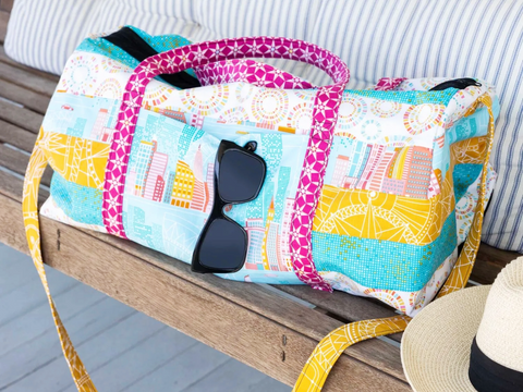 A colorful patchwork weekender bag with city and geometric patterns sits on a wooden bench.