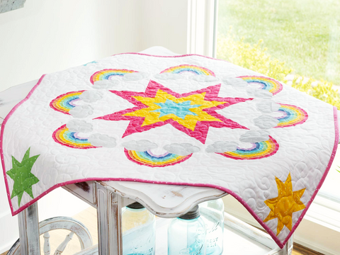 A white quilt with a large pink, yellow, and blue star surrounded by rainbows and clouds.