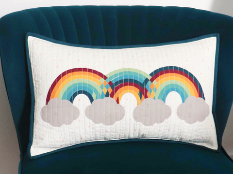 A rectangular white pillow with three colorful rainbows and gray clouds rests on a dark teal chair.
