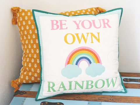 A white pillow with "BE YOUR OWN RAINBOW" text and a rainbow graphic.