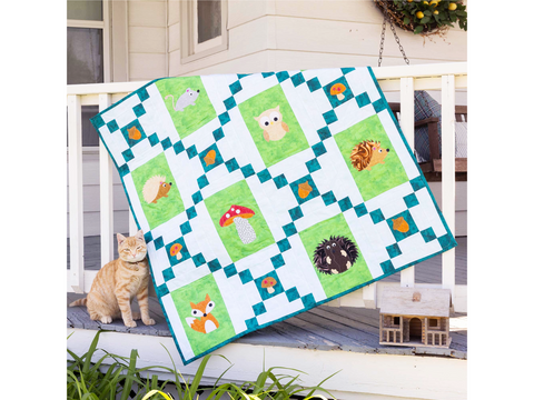 A quilt with forest animal and mushroom appliques hangs on a porch railing next to a cat.