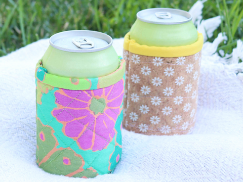 Two patterned can cozies hold light green cans on a white blanket with grass in the background.
