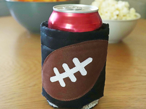 A red soda can sits in a black can cozy with a brown football applique.