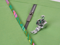 An adjustable zipper and piping foot is shown on green fabric with a zipper and colorful piping.