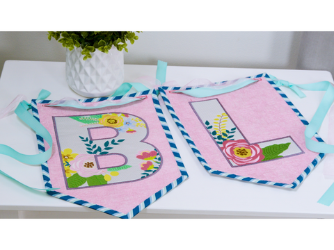 Two pink fabric banners, one with a floral "B" and one with a floral "L," tied with ribbons.