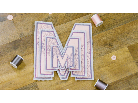 A stack of embroidered "M" appliques with floral designs, thread spools, and buttons on a wooden surface.