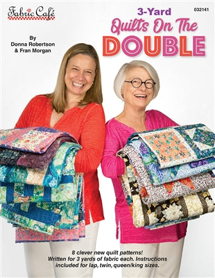 Two smiling women hold stacks of colorful quilts.