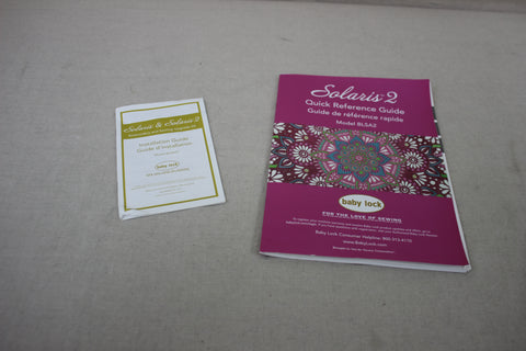 Two Baby Lock Solaris 2 manuals, one white and one magenta, lie on a gray surface.