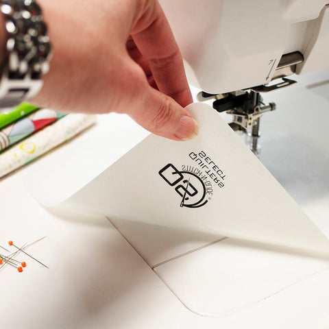 A hand places a white Stitch N Glide sheet with a black logo on a sewing machine.