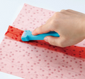 A hand uses a blue Clover Roll & Press tool to flatten a red fabric seam on a pink patterned fabric.