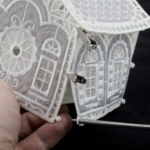 A hand holds a white lace house, with metal clips securing its roof and walls.